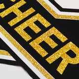 Close-up detail of gold glitter "CHEER" lettering and striped trim on a black and white cheerleading uniform top.